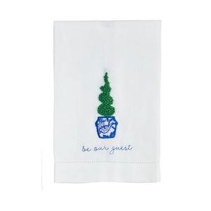 A white tea towel with a boxwood plant in a blue and white pot with "be our guest" embroidered below.