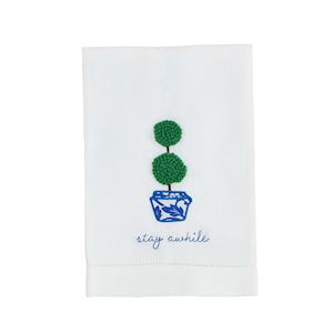 A white tea towel with a boxwood plant in a blue and white pot with "stay awhile" embroidered below.