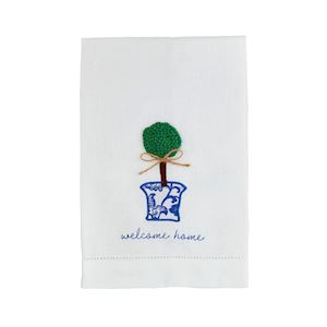 A white tea towel with a boxwood plant in a blue and white pot with "welcome home" embroidered below.
