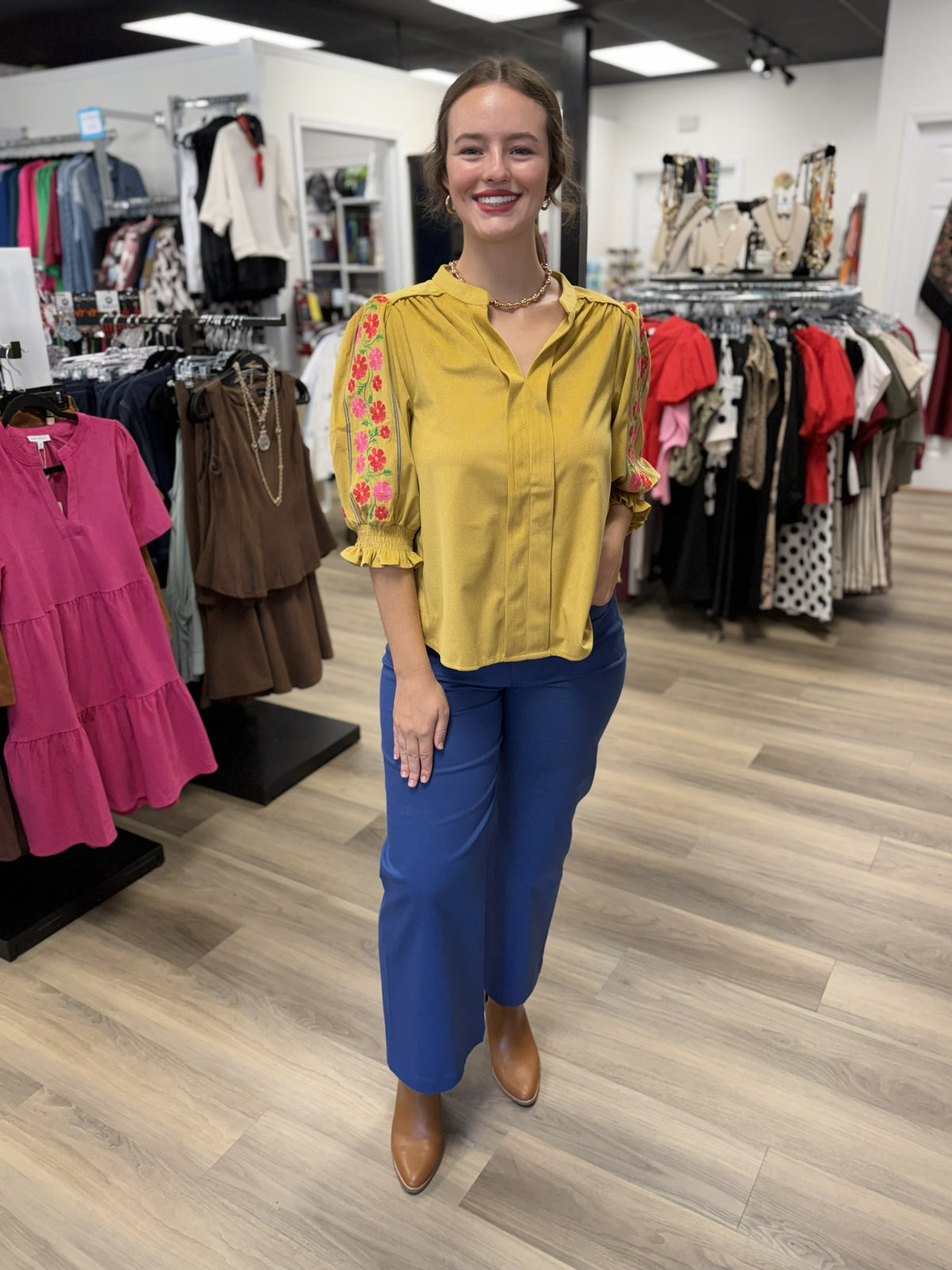 This is a mustard yellow blouse with a relaxed, feminine silhouette and subtle sheen. It features a split V-neckline with a banded collar, soft gathering at the shoulders, and a clean front placket. The three-quarter puff sleeves are the standout detail, featuring colorful floral embroidery in pink, coral, and green along the outer arms. Smocked cuffs with delicate ruffle trim complete the sleeves, giving the top a playful, polished finish.
