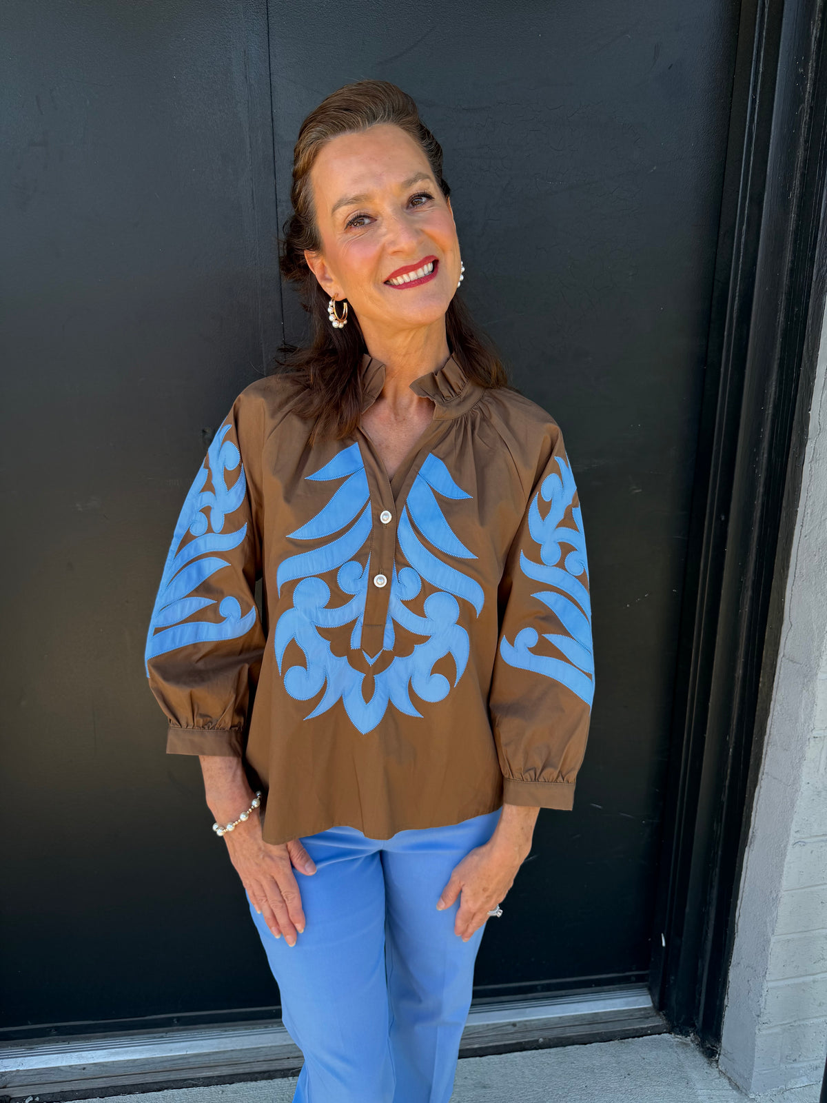 A woman wearing a brown button-up blouse with light blue scrollwork embroidery on the front and sleeves, paired with light blue pants, standing in front of a dark door.