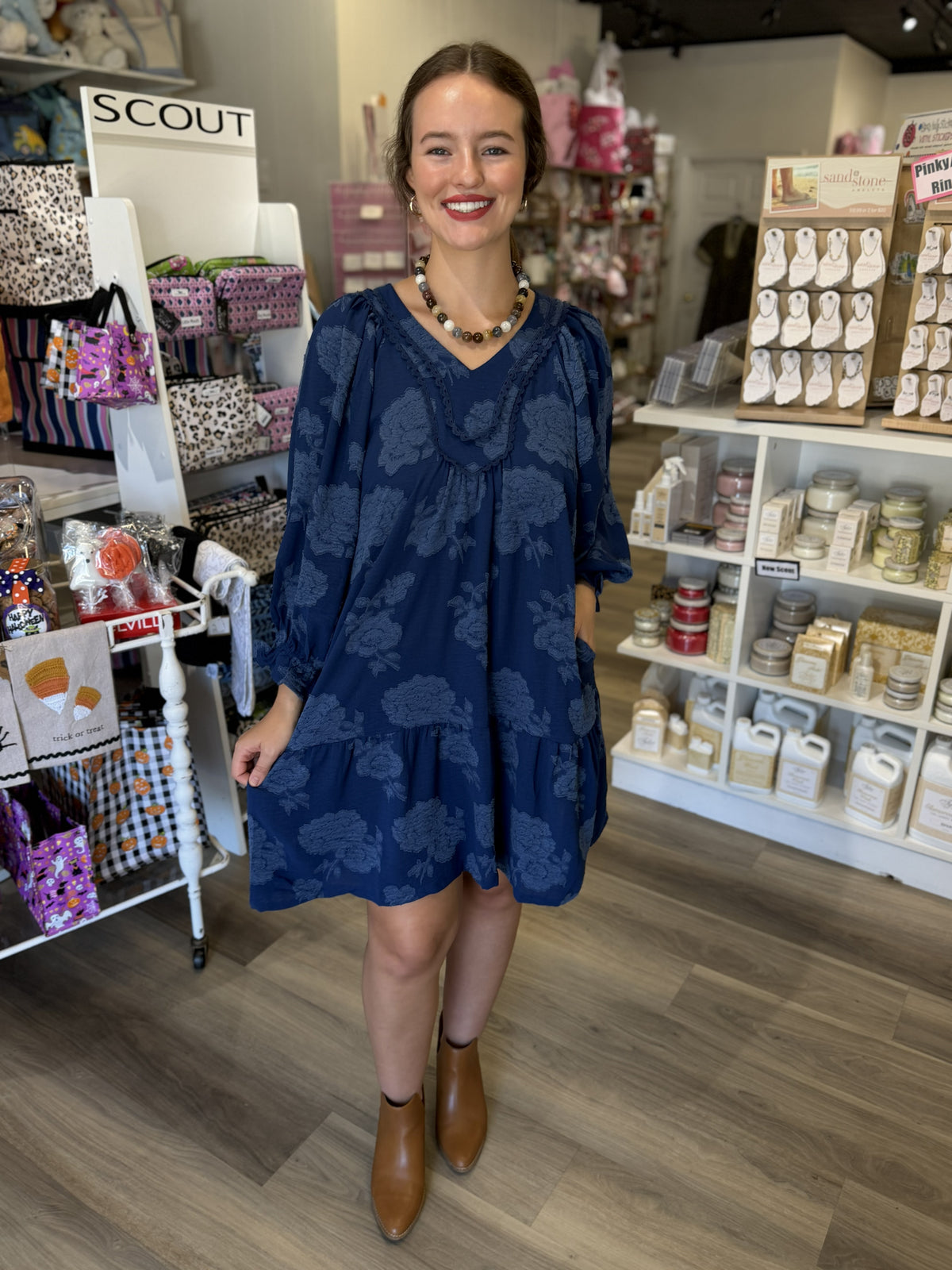 This is a navy blue floral dress with a relaxed, flowy silhouette and subtle tonal floral detailing throughout. It features a V-neckline framed with textured lace and ruffle trim, adding a feminine, romantic touch. The three-quarter-length balloon sleeves have gentle volume and gathered cuffs, while the loose bodice flows into a gathered tiered skirt that falls just above the knee. The monochromatic floral texture gives this easy-to-wear dress an elevated look, perfect for fall outings, church, dinner, or e