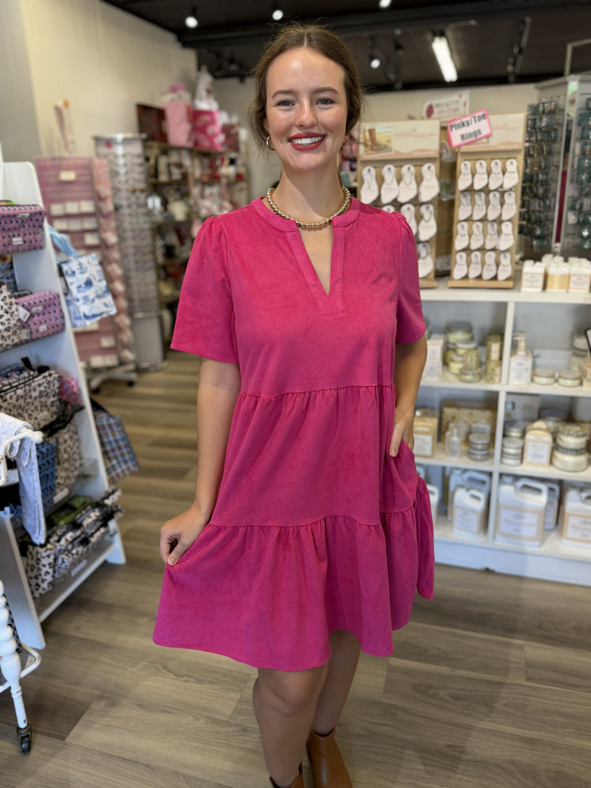 This is a vibrant fuchsia pink tiered dress with a relaxed, flowy silhouette. It features a split V-neckline, short sleeves with subtle gathering at the shoulders, and a loose-fitting bodice. The skirt is designed with two gathered tiers, creating soft volume and movement, and falls to around the knee. The lightweight, softly textured fabric and easy fit give it a polished yet playful look that can be dressed up with accessories or styled casually for everyday wear.