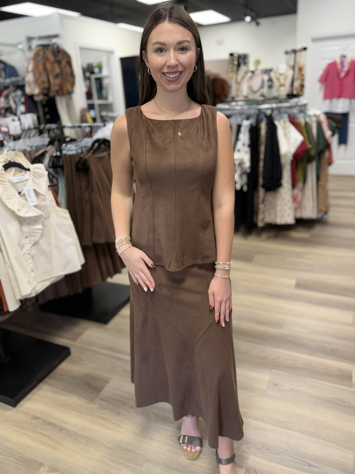 This outfit is a coordinating chocolate-brown two-piece set with a soft, suede-like finish. The sleeveless top features a square neckline, wide straps, vertical seam detailing, a fitted bodice, and a subtly flared hem. It’s paired with a matching high-waisted midi/maxi skirt that has a streamlined fit through the hips and gently flares toward the hem, with curved seam detailing adding shape and interest.