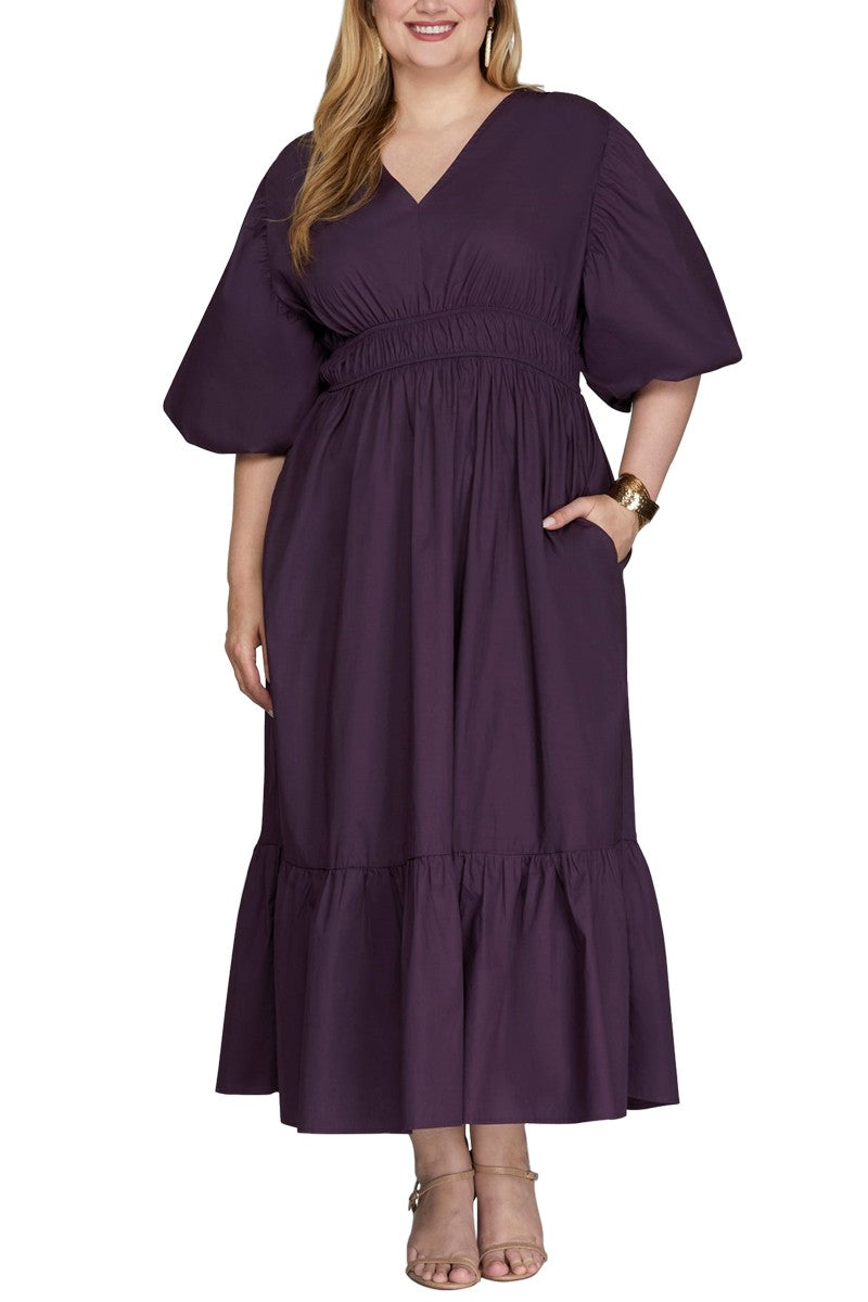 The dress is a deep purple, midi-length, with a V-neckline and short, wide sleeves that drape around the mid-upper arm. The waist has an elastic band with the skirt flares out with a ruffled hem.