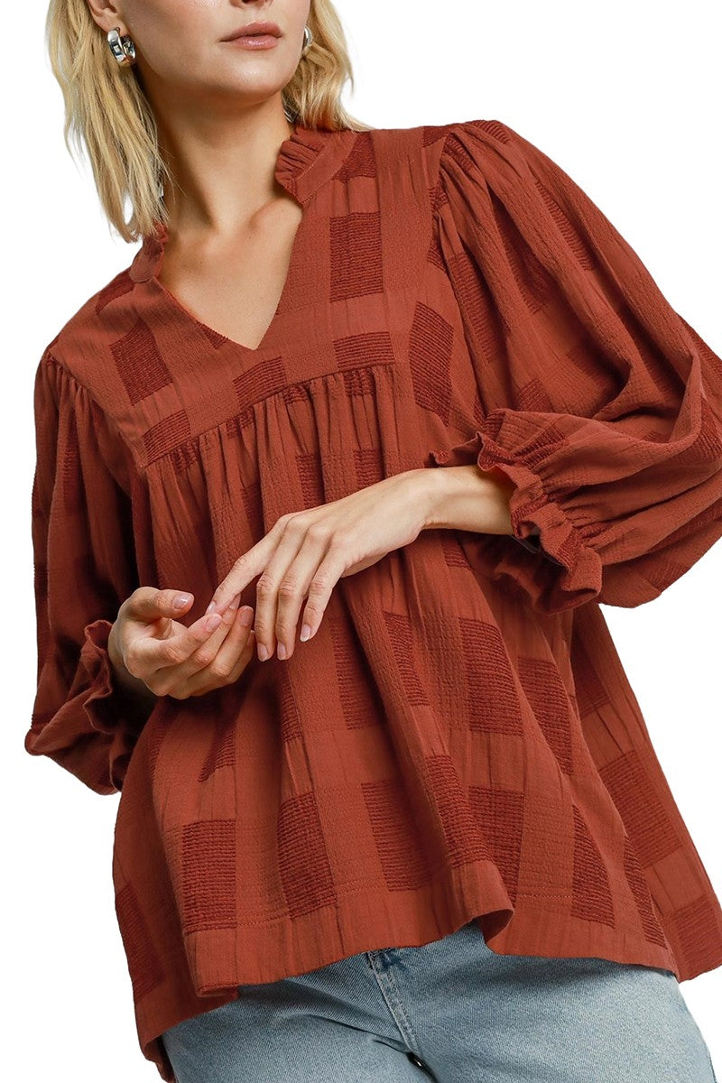 A long-sleeved rust colored blouse with a checkered pattern. It features a V-neckline and gathered detailing at the front, creating a loose, flowy fit.