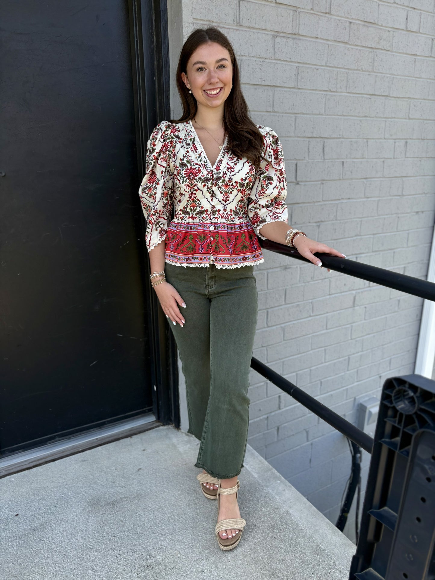 This top is a romantic, vintage-inspired blouse with a detailed, feminine design:

Ivory/cream base with an intricate red, burgundy, olive, and green floral/ornamental print
V-neckline with delicate lace-like trim
Long sleeves with gently gathered, puffed shoulders and fitted cuffs
Button-front closure through the bodice
Defined waist with a flattering, slightly fitted shape
A striking red printed border along the lower portion
Scalloped lace trim along the hem and sleeve cuffs
Slightly peplum-like shape at