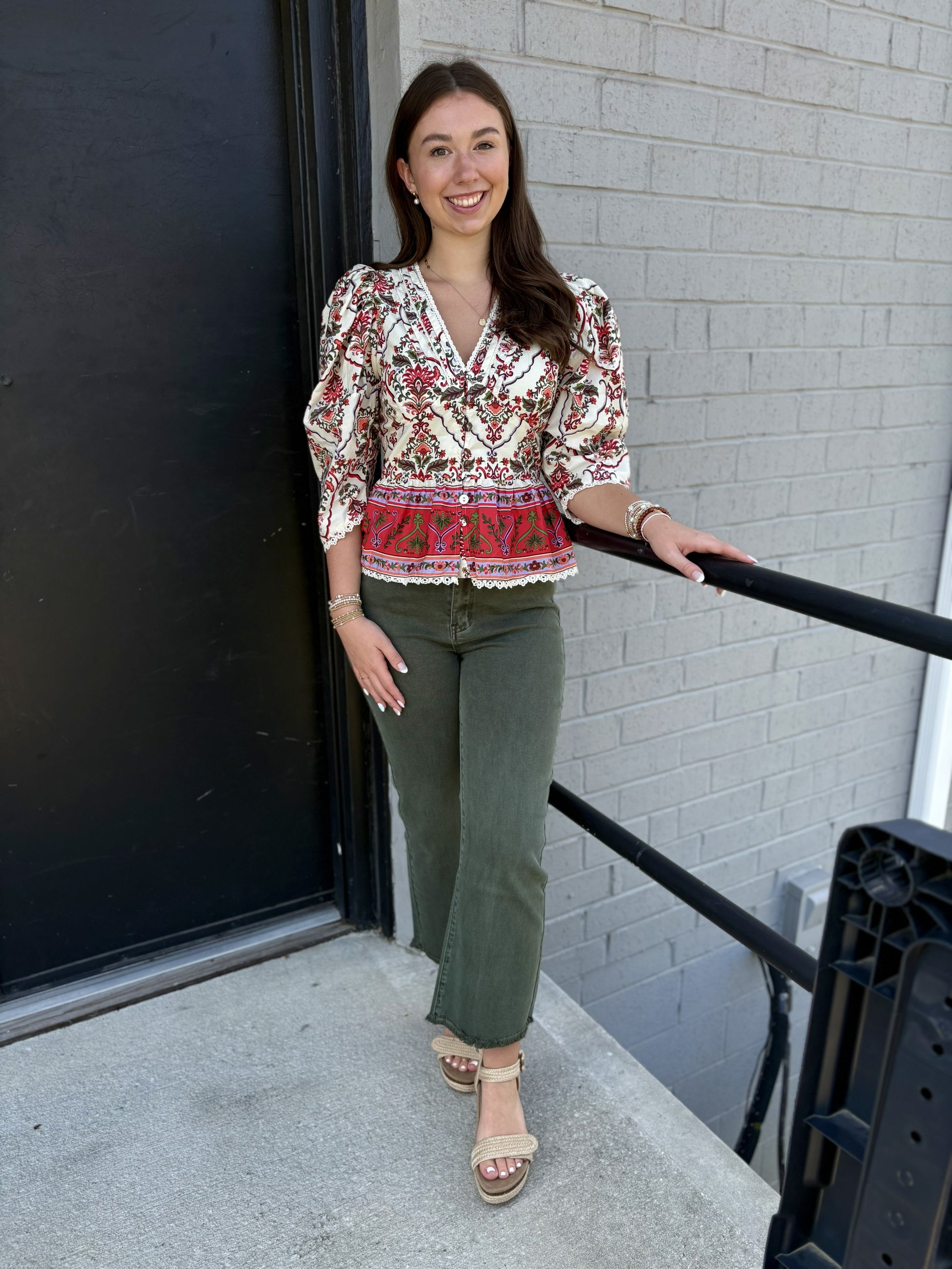 This top is a romantic, vintage-inspired blouse with a detailed, feminine design:

Ivory/cream base with an intricate red, burgundy, olive, and green floral/ornamental print
V-neckline with delicate lace-like trim
Long sleeves with gently gathered, puffed shoulders and fitted cuffs
Button-front closure through the bodice
Defined waist with a flattering, slightly fitted shape
A striking red printed border along the lower portion
Scalloped lace trim along the hem and sleeve cuffs
Slightly peplum-like shape at