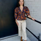 This top has a rich, bohemian-inspired look with beautiful vintage detailing:

Deep navy blue background with an intricate rust, copper, and warm gold floral paisley print
Deep V-neckline accented with delicate lace-like navy trim
Button-front design with covered buttons down the center
Three-quarter sleeves with softly gathered shoulders for a subtle puffed effect
Smocked/gathered waistline that creates a flattering, defined shape
Peplum-style hem that gently flares away from the waist
Decorative scalloped