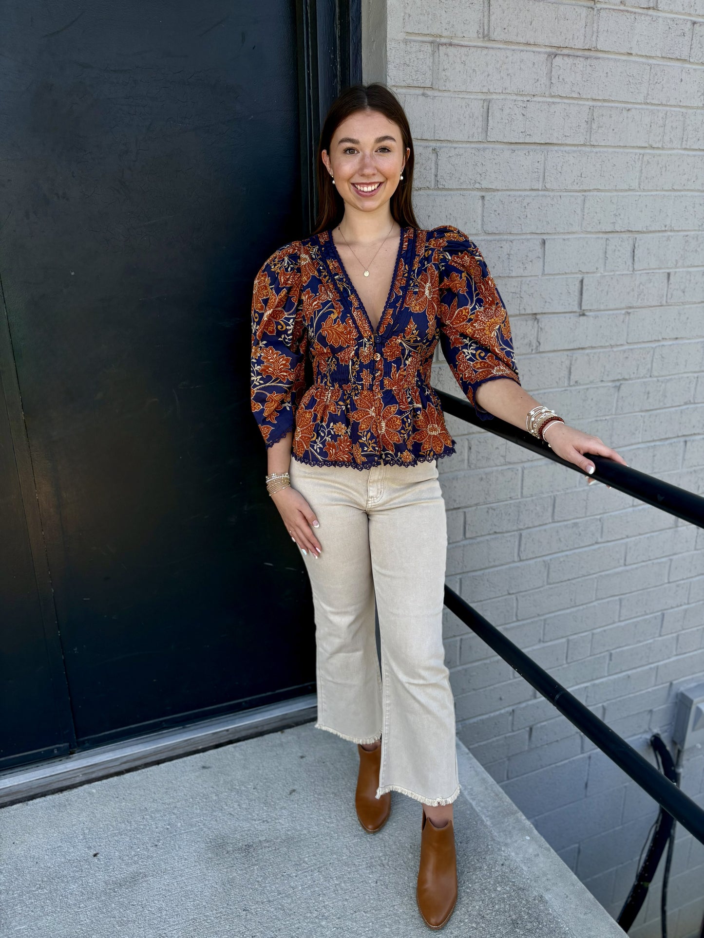 This top has a rich, bohemian-inspired look with beautiful vintage detailing:

Deep navy blue background with an intricate rust, copper, and warm gold floral paisley print
Deep V-neckline accented with delicate lace-like navy trim
Button-front design with covered buttons down the center
Three-quarter sleeves with softly gathered shoulders for a subtle puffed effect
Smocked/gathered waistline that creates a flattering, defined shape
Peplum-style hem that gently flares away from the waist
Decorative scalloped