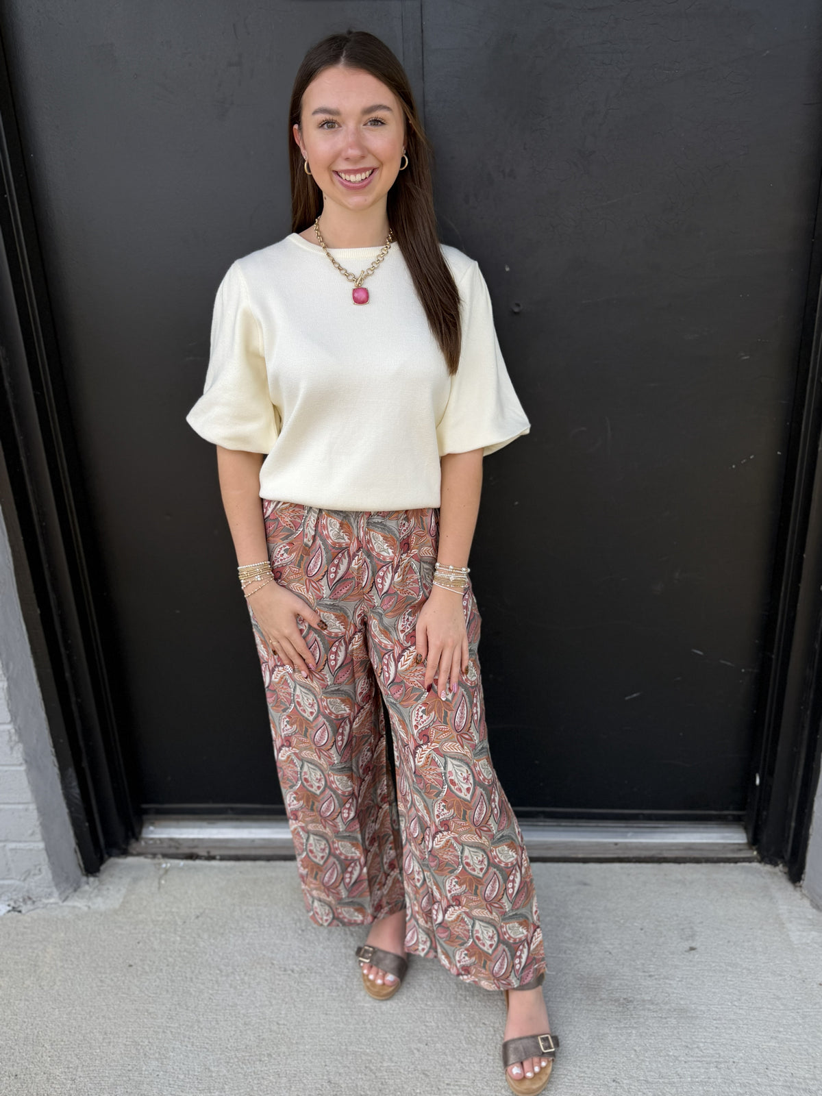 This effortlessly chic outfit pairs a soft ivory knit top with statement-making paisley wide-leg pants. The top features a classic round neckline, relaxed fit, and feminine elbow-length flutter sleeves for an easy, polished look. The flowy pants feature an intricate paisley print in warm rust, taupe, cream, and muted sage tones with a flattering high-rise waist and wide-leg silhouette. Finished with layered gold jewelry and neutral sandals, this outfit is perfect for work, lunch dates, or an elevated everyd