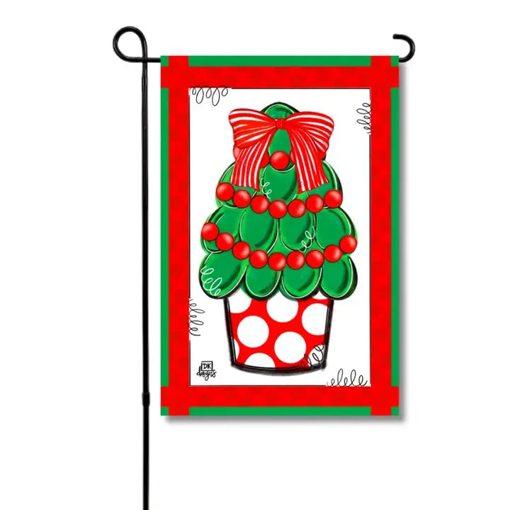 Christmas garden flag featuring a whimsical green Christmas tree decorated with red ornaments and garland, topped with a large red bow and displayed in a red-and-white polka dot planter.