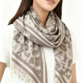 Leopard-print scarf in soft gray and beige tones, featuring striped accents, a subtle floral leopard pattern, and fringed edges. The scarf is draped around a woman’s neck over a white top.