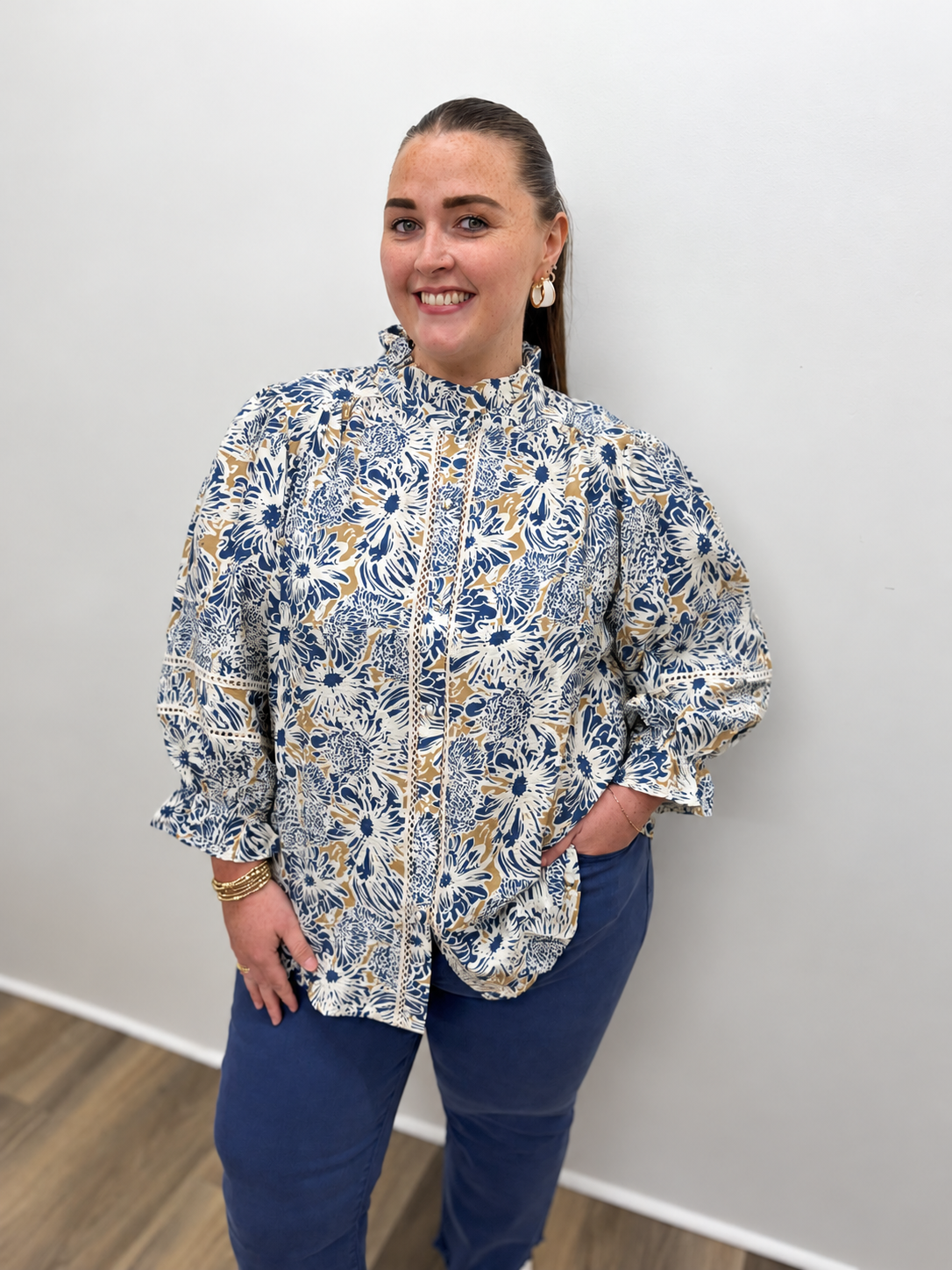 Women’s blue, cream, and tan floral print blouse featuring a ruffled high neckline, button-front design, three-quarter puff sleeves, and delicate lace trim details.
