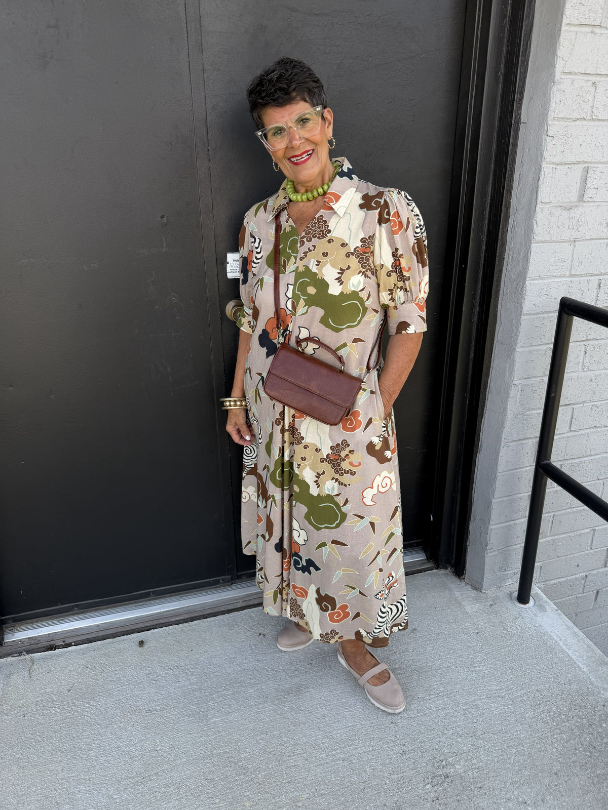 This is a beautiful vintage-inspired maxi dress with an artistic botanical and animal print, combining relaxed elegance with a playful, statement-making pattern.

Color: Soft taupe/beige background accented with olive green, rust, cream, chocolate brown, black, and touches of coral.
Print: An intricate nature-inspired print featuring abstract flowers, foliage, organic shapes, and whimsical animal motifs, including zebra-like figures.
Neckline: Classic shirt collar with a partially open button-front neckline