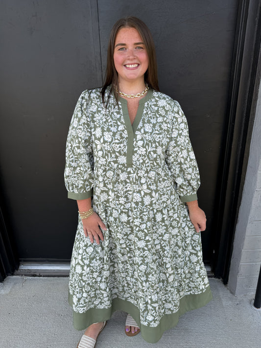 This is a sage green floral maxi dress featuring an allover white botanical print for a fresh, feminine look. It has a split V-neckline with solid sage trim, along with softly puffed three-quarter sleeves finished with coordinating cuffs. The relaxed bodice flows into a tiered, gathered skirt, creating an airy, flattering silhouette. A wide solid sage border at the hem adds a polished contrast and ties the design together. The flowy maxi length makes it a beautiful option for church, brunch, showers, family