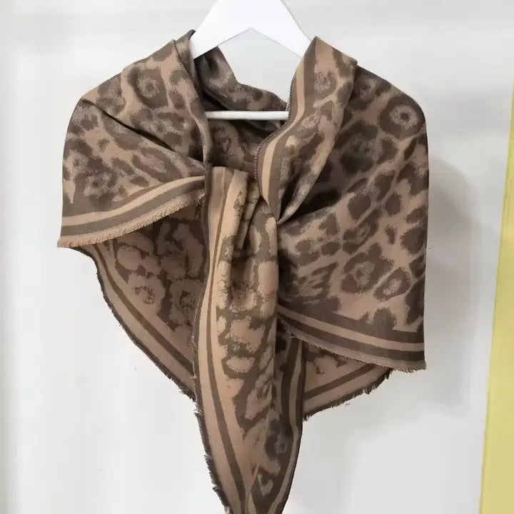 Leopard-print scarf in neutral taupe, brown, and beige tones, featuring a striped border and a knotted center. Displayed on a white hanger against a light background.
