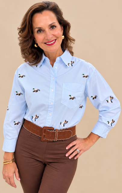 A woman modeling a light blue western-style button-down shirt with embroidered birds, paired with brown pants and a brown belt, against a solid beige background.