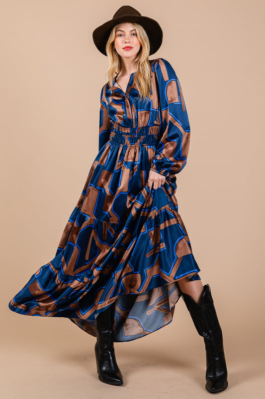 A long-sleeved, full-length dress with a flowing skirt. A bold geometric pattern with rectangles and lines in shades of blue, brown, and beige. The dress has a cinched waist, giving it a slight balloon effect on the top. A V-neckline style, with cuffed sl