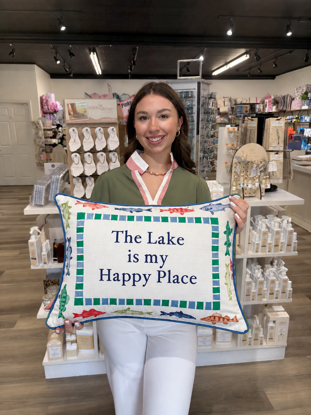This decorative rectangular lake-themed pillow features the phrase “The Lake is my Happy Place” in bold navy lettering across a crisp cream background. A colorful border of fish motifs in shades of blue, green, red, and brown surrounds the design, accented by a checkered blue-and-green inner border. Finished with bright blue piping, this playful pillow is perfect for a lake house, cabin, porch, or coastal-inspired space.