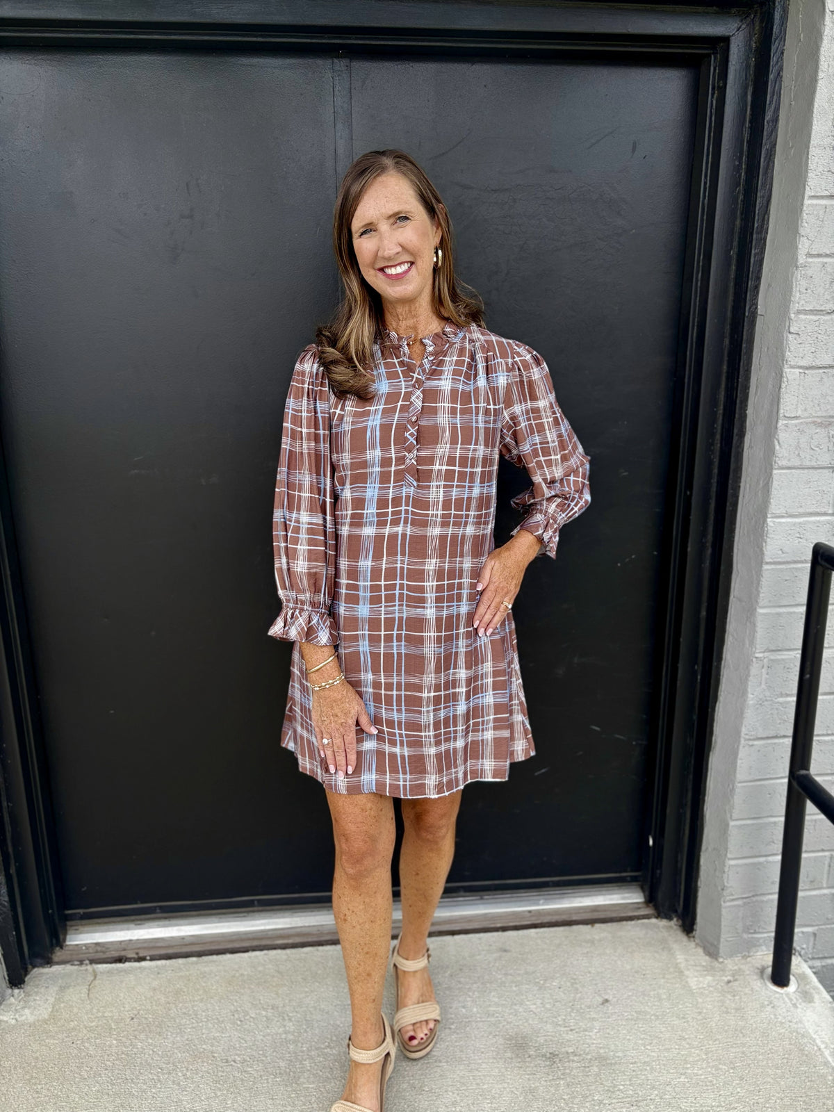 This dress has a pretty, relaxed boutique style with a subtle bohemian feel:

Warm chocolate-brown base with an allover white and light-blue windowpane plaid
High, softly gathered neckline with a small tie detail at the center
Partial button-front placket with delicate decorative stitching
Long, slightly sheer-looking sleeves with gathered cuffs and feminine ruffle-like trim
Relaxed, A-line silhouette that falls loosely from the body
Short, above-the-knee length for an easy, flattering shape
Lightweight-loo