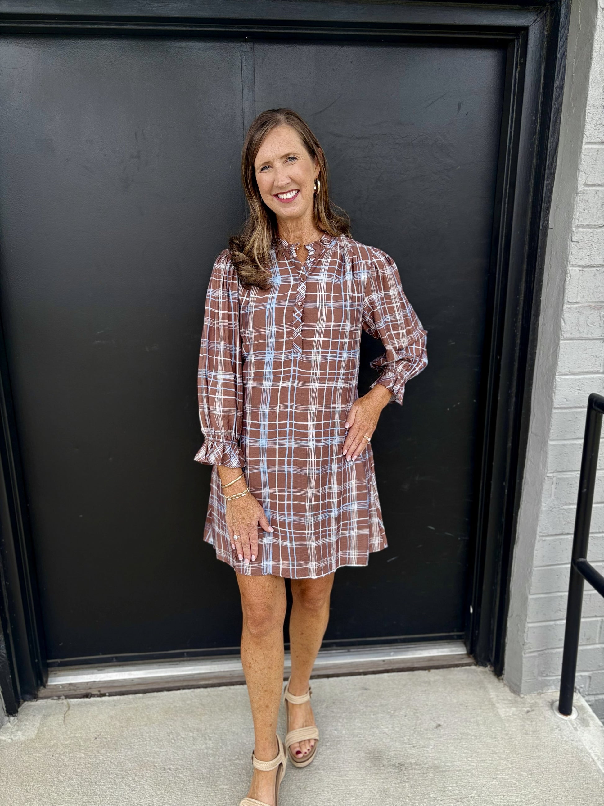 This dress has a pretty, relaxed boutique style with a subtle bohemian feel:

Warm chocolate-brown base with an allover white and light-blue windowpane plaid
High, softly gathered neckline with a small tie detail at the center
Partial button-front placket with delicate decorative stitching
Long, slightly sheer-looking sleeves with gathered cuffs and feminine ruffle-like trim
Relaxed, A-line silhouette that falls loosely from the body
Short, above-the-knee length for an easy, flattering shape
Lightweight-loo