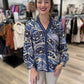 This is a flowy blue mixed-print blouse featuring an abstract combination of animal-inspired spots and swirling botanical patterns in navy, slate blue, periwinkle, cream, and black. It has a deep V-neckline with contrasting navy striped trim that extends down the center front, along with a subtle ruffled stand collar. The long, billowy sleeves finish with gathered cuffs, while the relaxed silhouette creates an easy, feminine fit.