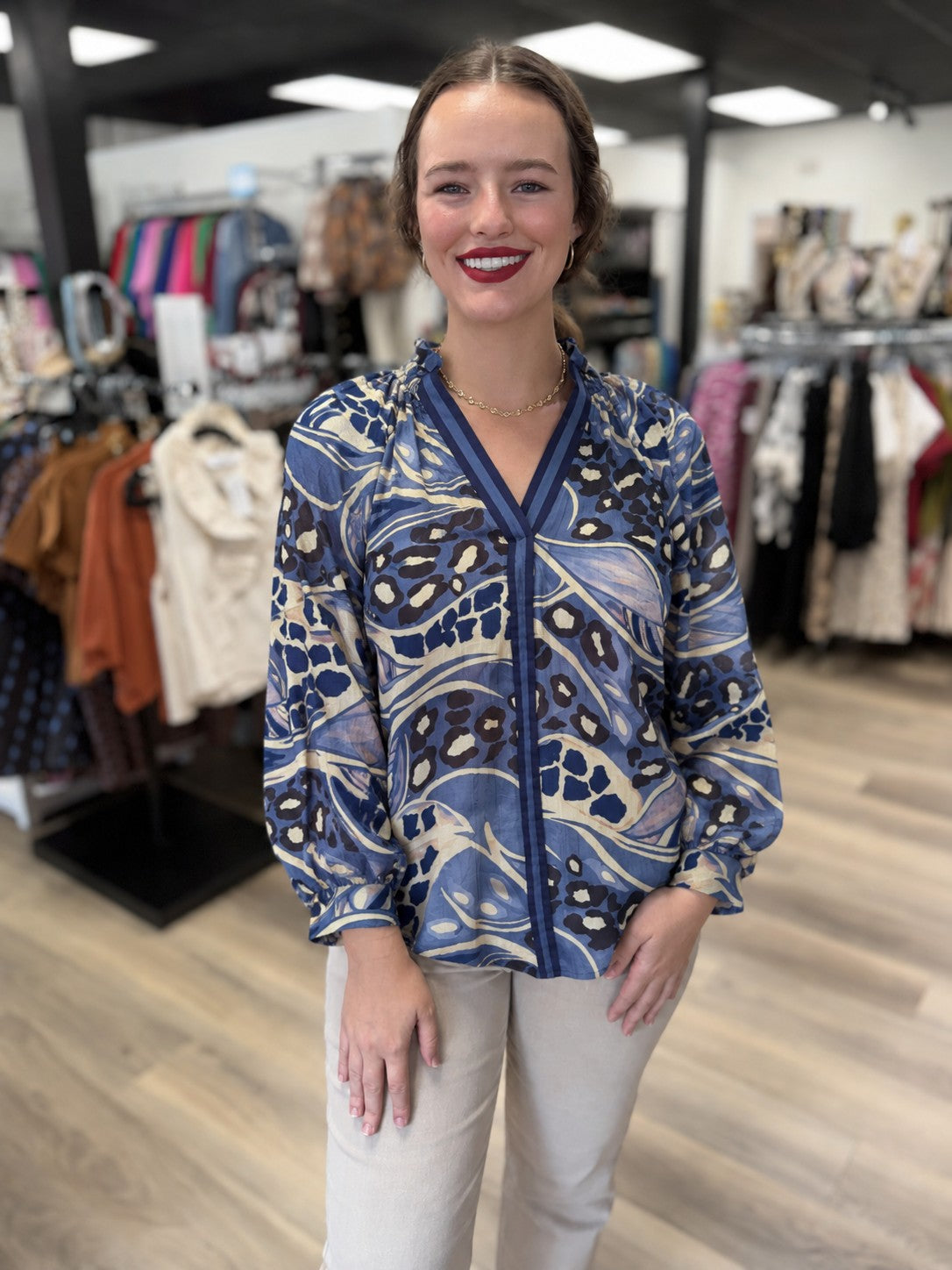 This is a flowy blue mixed-print blouse featuring an abstract combination of animal-inspired spots and swirling botanical patterns in navy, slate blue, periwinkle, cream, and black. It has a deep V-neckline with contrasting navy striped trim that extends down the center front, along with a subtle ruffled stand collar. The long, billowy sleeves finish with gathered cuffs, while the relaxed silhouette creates an easy, feminine fit.