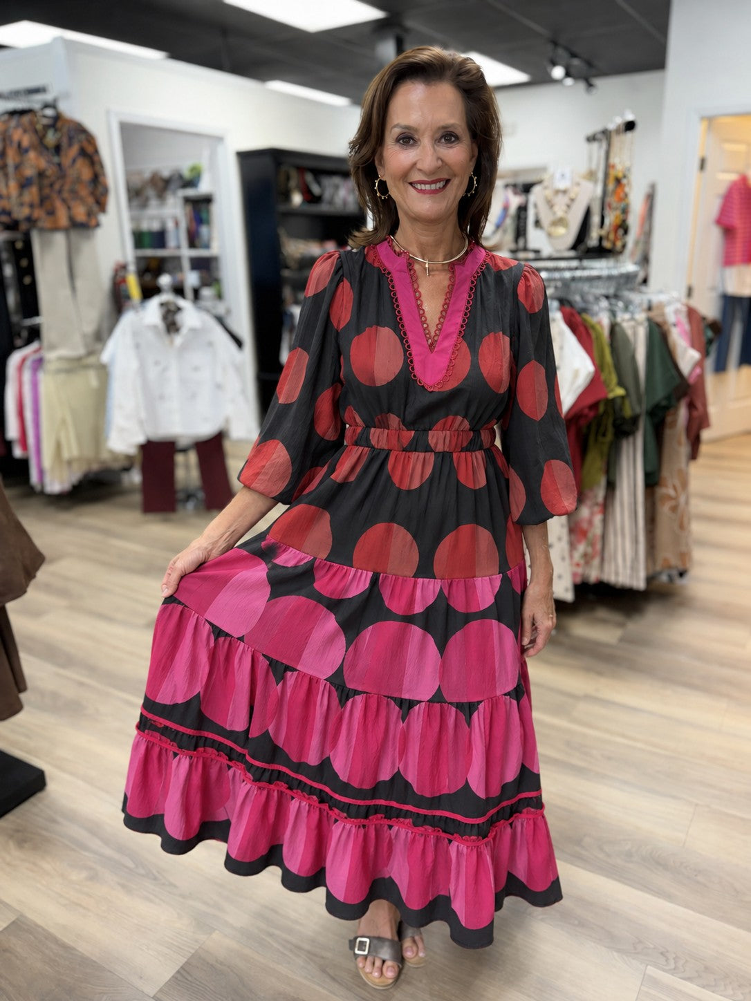 This is a bold black and hot-pink maxi dress with a playful oversized polka-dot design. The bodice features rusty coral dots against a black background, a deep V-split neckline with bright pink contrast trim and decorative loop detailing, and relaxed three-quarter-length puff sleeves.

A gathered elastic waist defines the silhouette before flowing into a full, tiered maxi skirt. The skirt transitions into large hot-pink circular motifs on black, with multiple gathered tiers and decorative pink trim near the