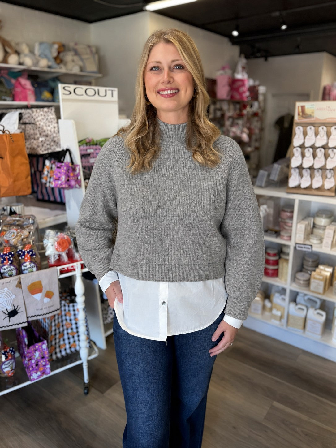 This top is a heather gray layered sweater-and-shirt style with a polished, preppy look. It features a soft ribbed knit sweater with a mock neckline, long sleeves, relaxed fit, and slightly cropped hem. A crisp white button-down layer extends beneath the sweater, creating a curved shirttail hem, with matching white cuffs peeking out at the wrists. The contrasting layers give it the look of a perfectly styled sweater over a classic white shirt in one easy piece.