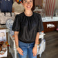 This is a black short-sleeve top with a subtle sheen and a relaxed, feminine silhouette. It features a round crew neckline with soft gathering across the front and gently puffed short sleeves with gathered shoulders. The slightly blouson-style bodice is finished with an elasticized hem and delicate ruffle trim, creating a flattering shape at the waist. The sleek black finish makes it an easy, versatile piece to dress up with trousers or skirts or wear casually with denim.