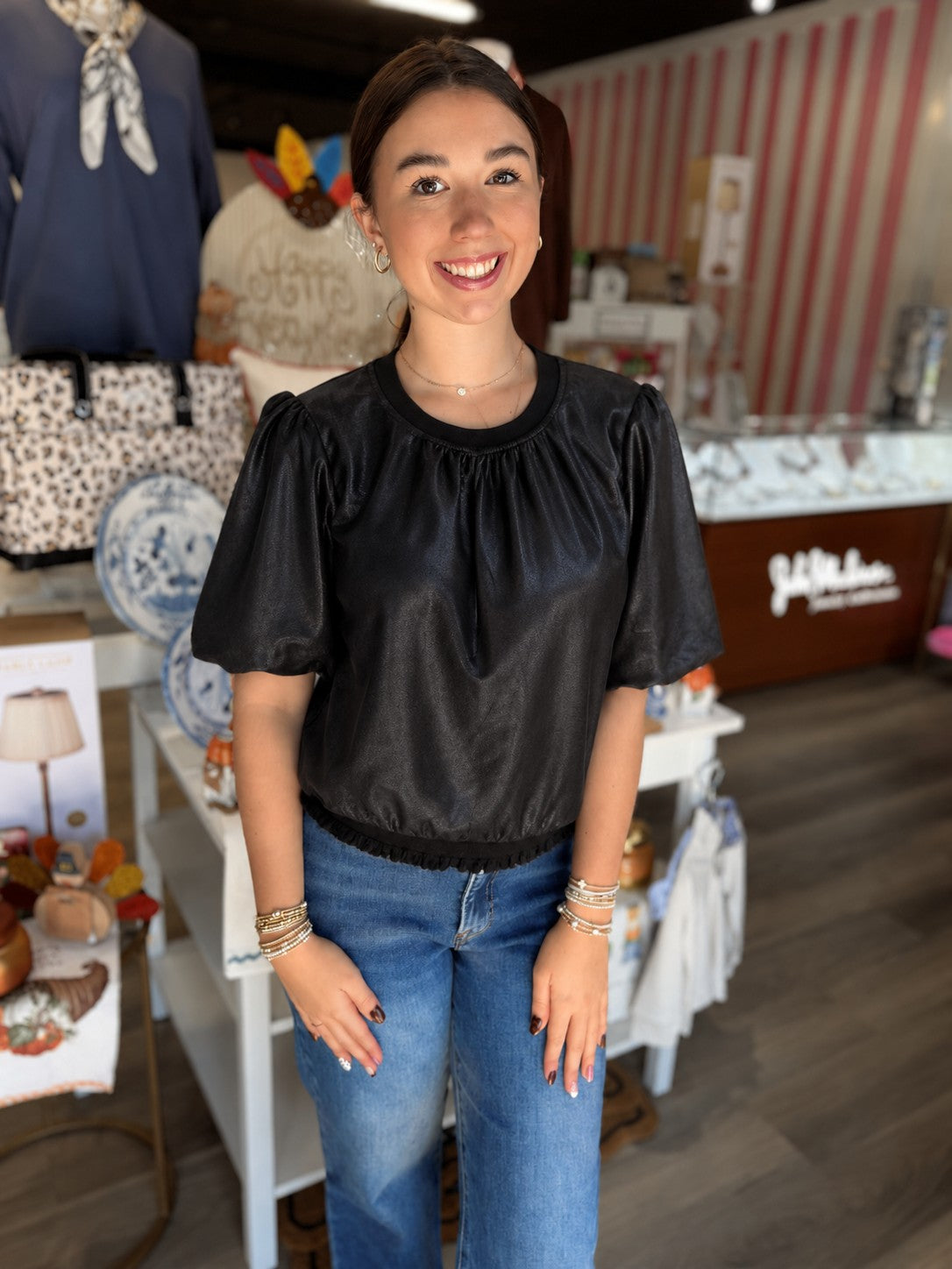 This is a black short-sleeve top with a subtle sheen and a relaxed, feminine silhouette. It features a round crew neckline with soft gathering across the front and gently puffed short sleeves with gathered shoulders. The slightly blouson-style bodice is finished with an elasticized hem and delicate ruffle trim, creating a flattering shape at the waist. The sleek black finish makes it an easy, versatile piece to dress up with trousers or skirts or wear casually with denim.