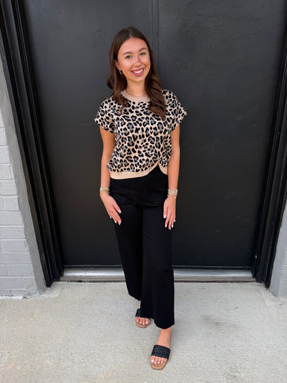 This is a chic leopard-print knit top with a feminine twisted-front detail and contrast trim.

Print: Classic leopard print in black, warm beige, and cream tones.
Neckline: Simple round crew neckline with a contrasting cream-colored ribbed edge.
Sleeves: Short, slightly relaxed cap sleeves, giving it an easy, polished shape.
Fit: Comfortable and gently fitted through the bodice without feeling overly tight.
Hem: The standout feature is the twisted/gathered center-front hem, creating a subtle sweetheart-like