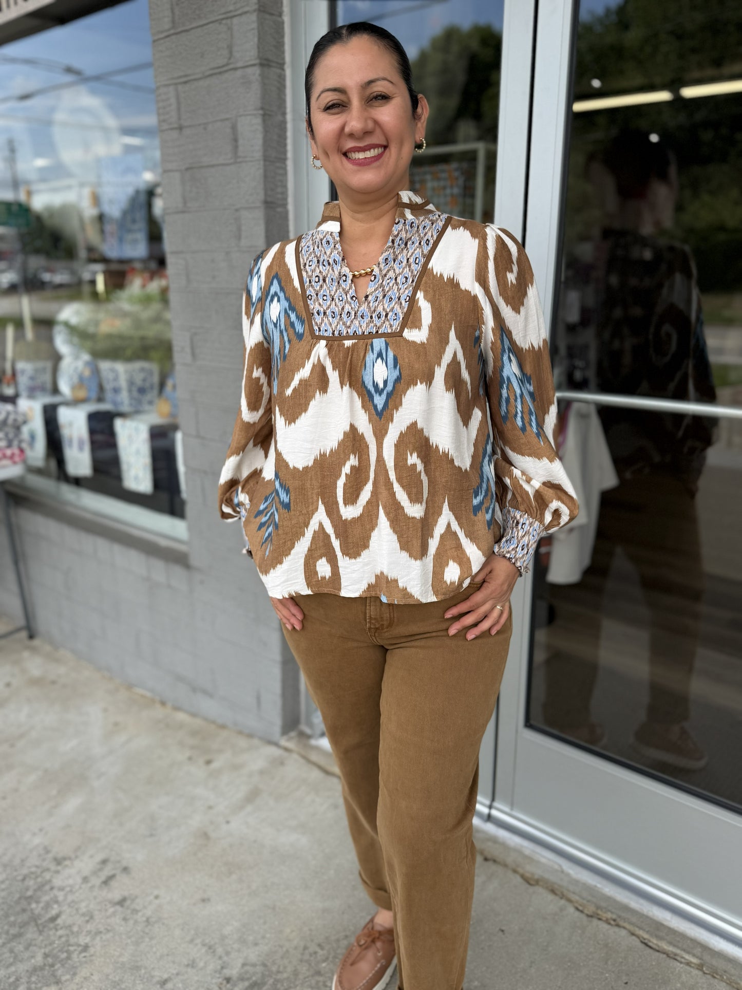 This bohemian-inspired printed blouse combines an earthy palette with beautiful statement details:

Color: Warm camel brown, ivory, and soft blue
Print: Bold, oversized geometric/ikat-inspired pattern throughout
Neckline: Split neckline with a structured, embroidered-looking bib panel in blue, ivory, and brown
Sleeves: Long sleeves with a relaxed, slightly voluminous shape and gathered cuffs
Cuffs: Coordinating patterned cuffs with a gathered finish
Fit: Relaxed, easy silhouette with a softly flowing body
H