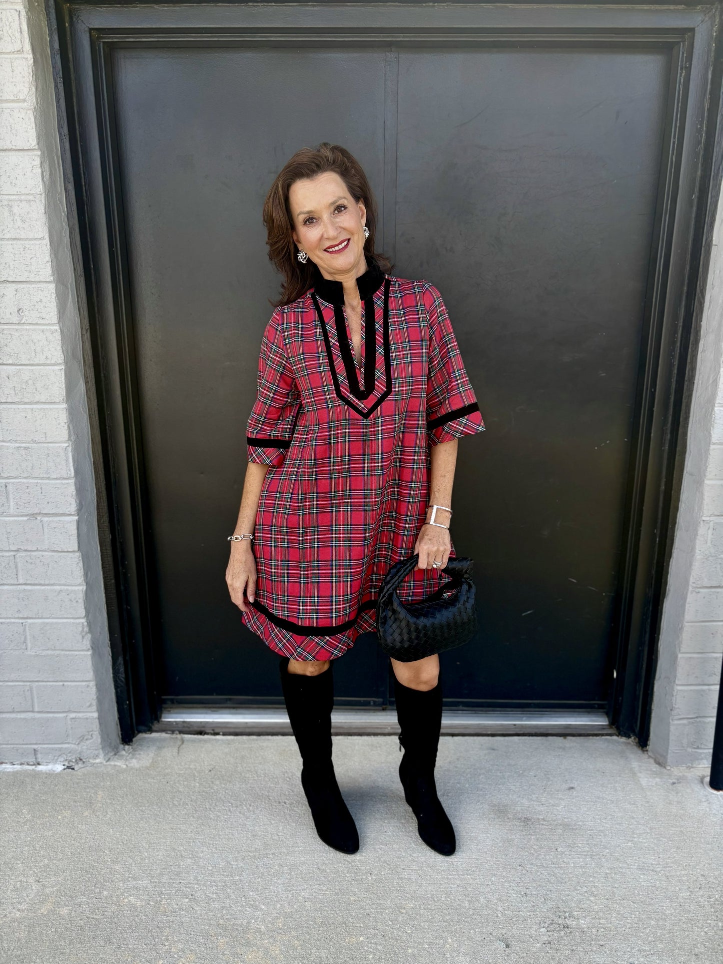 This dress has a bold, classic-meets-modern plaid look:

Red tartan plaid pattern with intersecting black, white, and subtle green lines
Relaxed, straight silhouette with a flattering, easy fit
V-neckline framed with contrasting black velvet-like trim
Black stand collar for a polished, sophisticated touch
Three-quarter sleeves finished with coordinating black trim
Black decorative detailing continues along the hem for a cohesive finish
Above-the-knee length, giving it a chic, youthful feel
The overall style