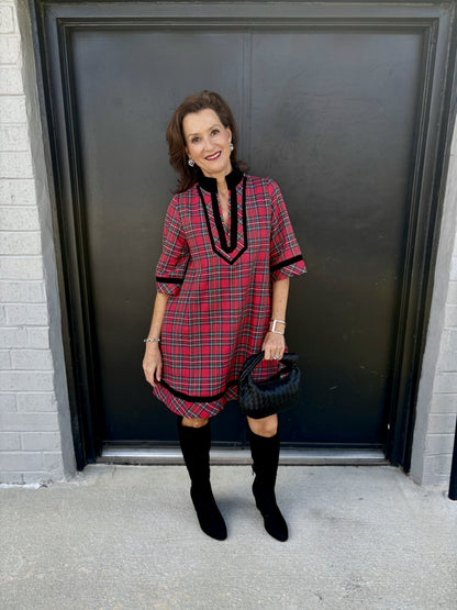 This dress has a bold, classic-meets-modern plaid look:

Red tartan plaid pattern with intersecting black, white, and subtle green lines
Relaxed, straight silhouette with a flattering, easy fit
V-neckline framed with contrasting black velvet-like trim
Black stand collar for a polished, sophisticated touch
Three-quarter sleeves finished with coordinating black trim
Black decorative detailing continues along the hem for a cohesive finish
Above-the-knee length, giving it a chic, youthful feel
The overall style