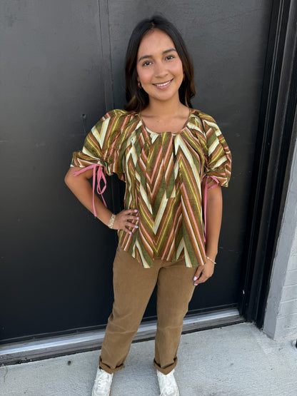 This top features a soft, flowing silhouette with a retro-inspired geometric print. It has:

Multicolor chevron/zigzag pattern in olive green, brown, blush pink, cream, and tan
Rounded scoop neckline with subtle gathering at the center
Short, voluminous sleeves with adjustable pink tie details at the cuffs
Relaxed, slightly flared fit that drapes away from the body
Lightweight, textured fabric that gives the top an airy, effortless feel
A slightly longer length that pairs easily with slim or straight-leg bo