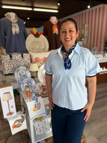 This top is a classic light-blue short-sleeve button-up blouse with a polished, preppy feel.

Soft pale blue color
Traditional pointed collar
Full button-front closure
Short, slightly puffed sleeves with gathered volume
Contrasting navy trim around the sleeve cuffs for a sporty, refined detail
Relaxed, straight silhouette with a slightly boxy fit
Lightweight woven fabric with a crisp, clean appearance
Styled with a navy patterned neck scarf, adding a sophisticated vintage-inspired touch