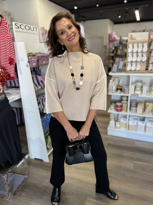 This is a soft beige knit top with a polished, modern silhouette. It features a high crew neckline, relaxed dropped shoulders, and elbow-length sleeves for an easy yet sophisticated look. The standout detail is the contrasting white piping that traces the shoulders and continues down the outer sleeves, adding a clean sporty-chic accent. The slightly cropped, boxy fit pairs beautifully with tailored pants or skirts.