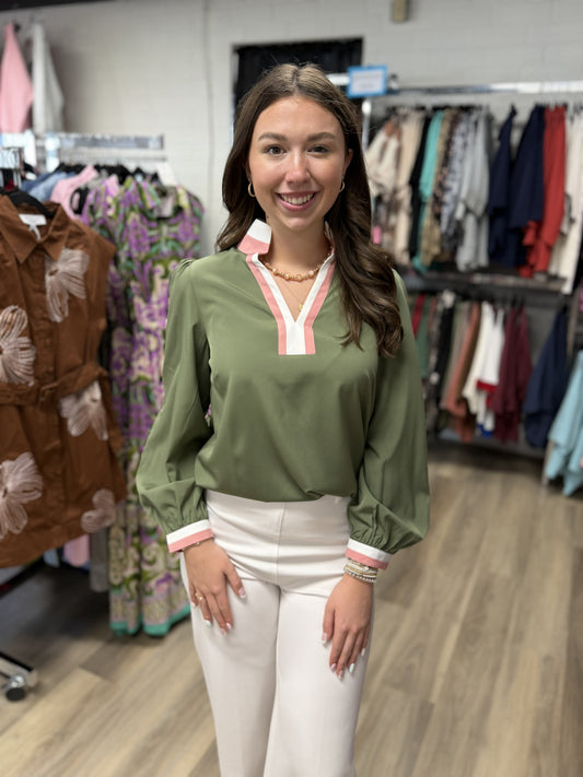 This is a soft olive-green blouse with a polished, preppy feel. It features a split V-neckline and stand collar accented with crisp white and blush-pink trim, giving the solid green body a fresh pop of color.

The blouse has a relaxed, flowy fit with long balloon sleeves, subtle gathering at the shoulders, and elasticized cuffs finished with matching pink-and-white trim. It’s an easy wear-to-work or everyday top that pairs beautifully with cream pants, denim, or tailored trousers.