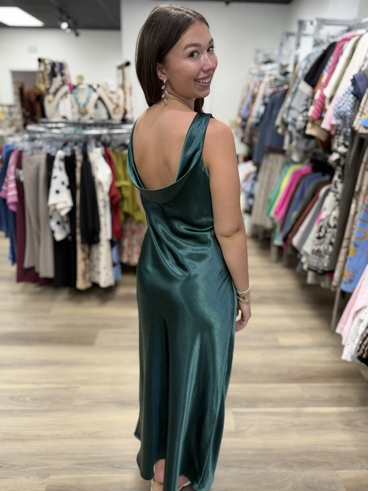 This is a rich emerald green satin maxi dress with an elegant, fluid silhouette. The lustrous fabric has a beautiful sheen that catches the light, giving the dress a sophisticated, dressy finish.

It features a sleeveless design with a softly rounded bateau neckline in front and a striking low scoop back with a draped cowl detail. The dress skims the figure before falling into a relaxed, flowing ankle-to-maxi length hem. Minimalist yet glamorous, it’s a beautiful option for weddings, formal events, holiday 