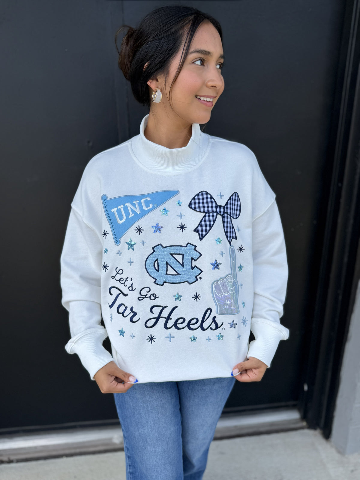 This is a white oversized mock-neck sweatshirt featuring a playful UNC Tar Heels–themed graphic across the front. The design combines Carolina blue, navy, and black details, including a UNC pennant, gingham bow, UNC logo, foam finger, stars, and “Let’s Go Tar Heels” lettering. Sparkly accents add a fun touch to the collegiate design. It has a ribbed mock neckline, dropped shoulders, long sleeves with ribbed cuffs, and a relaxed, roomy fit for a cozy game-day look.