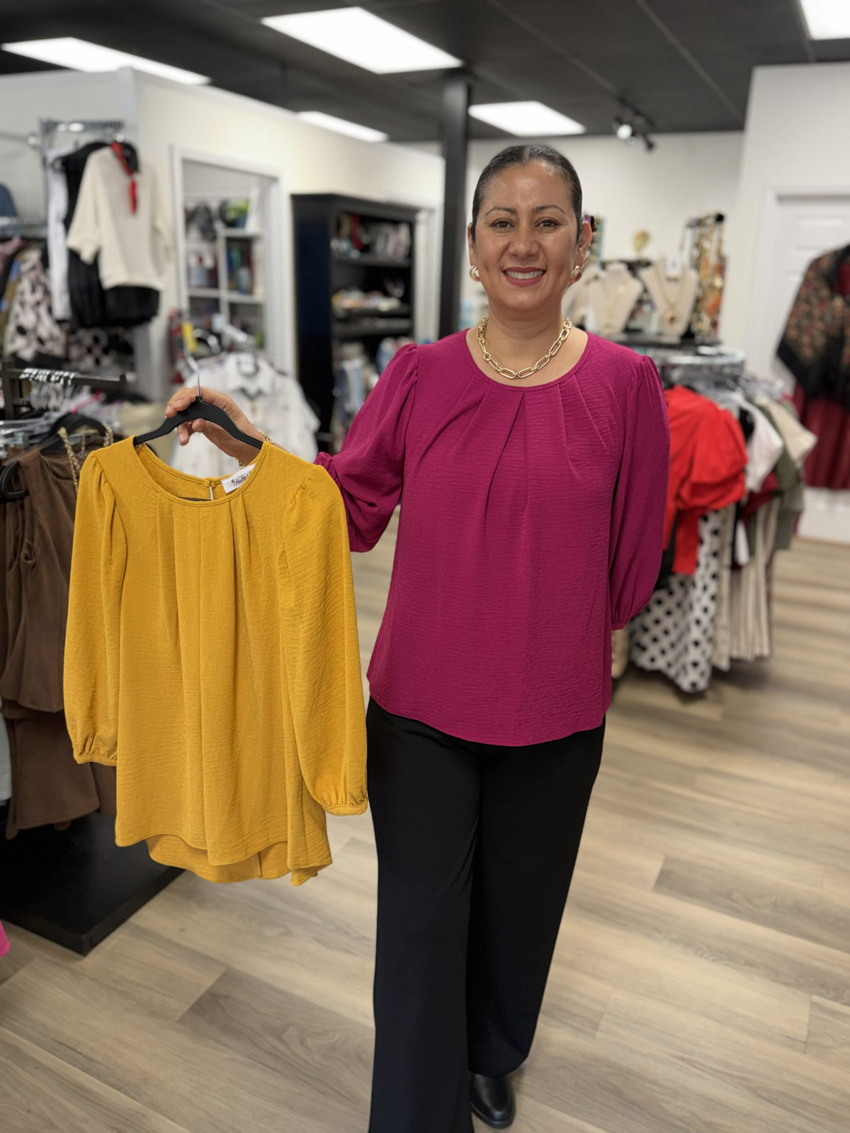 This is a rich magenta long-sleeve blouse with a softly textured finish and an easy, relaxed silhouette. It features a classic round neckline with delicate pleating across the front, adding subtle dimension and drape. The sleeves have gentle volume through the arms and are finished with elasticized cuffs for a softly gathered effect. The curved hem and flowy fit give it a polished yet comfortable look that pairs beautifully with black pants, denim, or skirts.