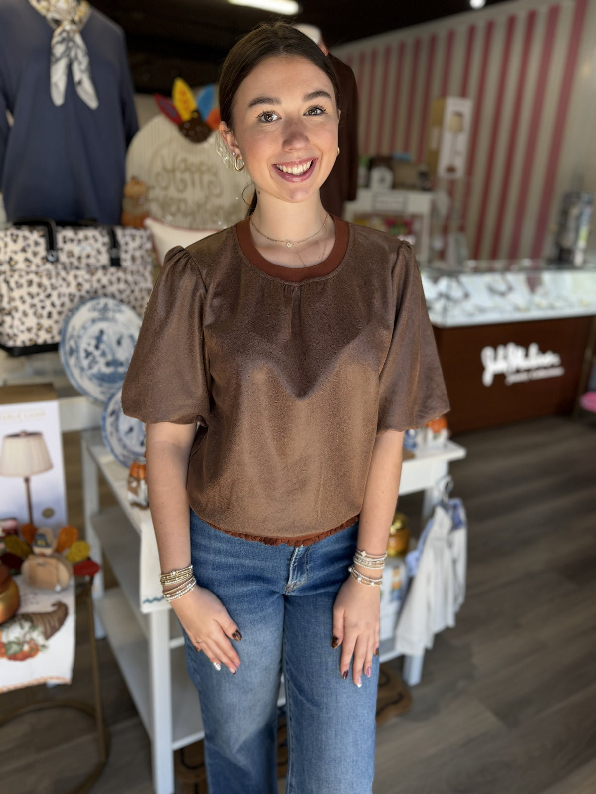 This is a rich chocolate brown short-sleeve top with a soft, suede-like texture and relaxed silhouette. It features a round neckline with contrasting rust-brown ribbed trim, subtle gathering across the front, and short puff sleeves with softly gathered shoulders for a feminine touch. A delicate ruffled trim along the hemline adds extra detail. The warm neutral color and textured finish make it an easy fall staple that pairs beautifully with denim, skirts, or trousers.