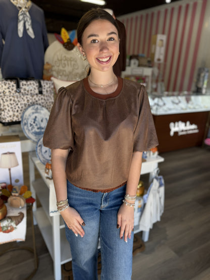 This is a rich chocolate brown short-sleeve top with a soft, suede-like texture and relaxed silhouette. It features a round neckline with contrasting rust-brown ribbed trim, subtle gathering across the front, and short puff sleeves with softly gathered shoulders for a feminine touch. A delicate ruffled trim along the hemline adds extra detail. The warm neutral color and textured finish make it an easy fall staple that pairs beautifully with denim, skirts, or trousers.