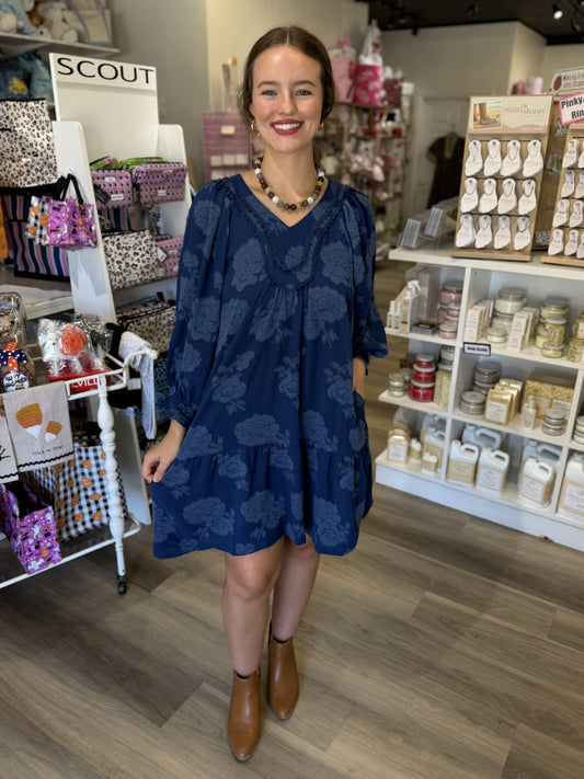 This is a navy blue floral dress with a relaxed, flowy silhouette and subtle tonal floral detailing throughout. It features a V-neckline framed with textured lace and ruffle trim, adding a feminine, romantic touch. The three-quarter-length balloon sleeves have gentle volume and gathered cuffs, while the loose bodice flows into a gathered tiered skirt that falls just above the knee. The monochromatic floral texture gives this easy-to-wear dress an elevated look, perfect for fall outings, church, dinner, or e