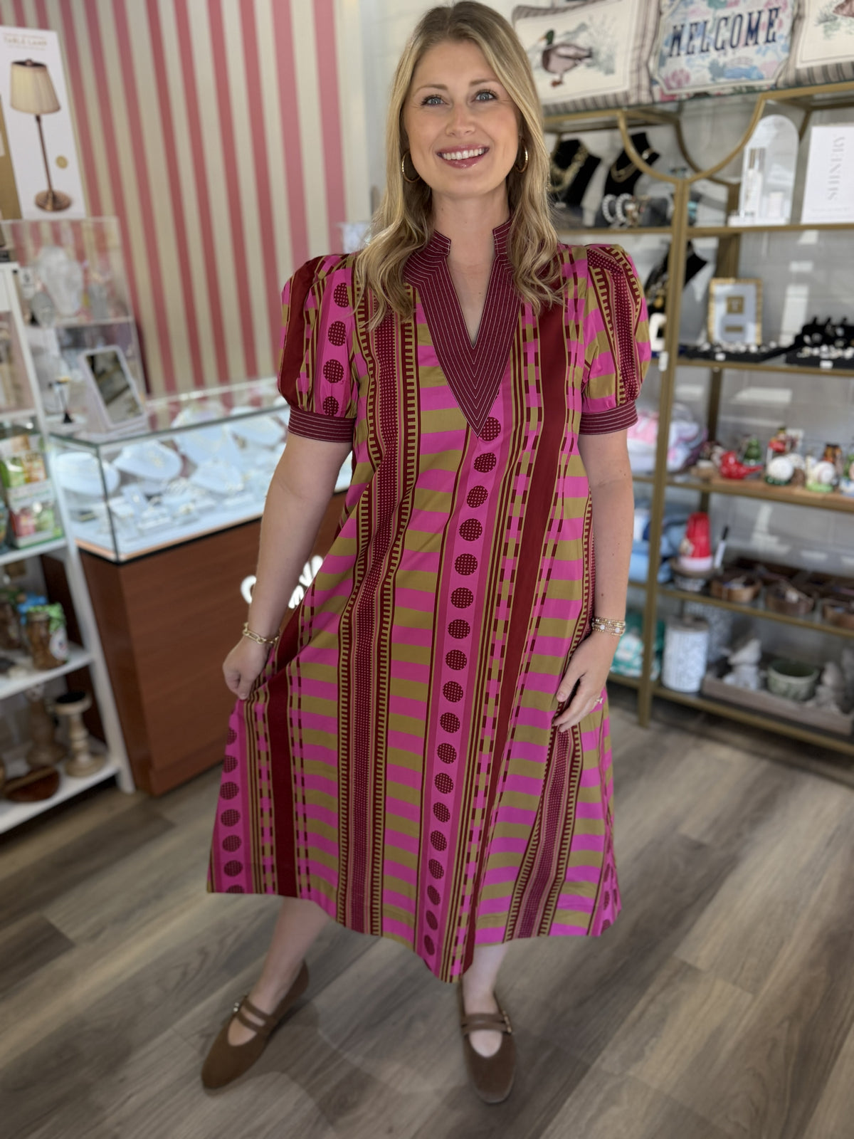 This is a bold pink, olive, and burgundy printed midi dress featuring a striking mix of stripes, geometric borders, and dotted motifs. It has a structured split V-neckline with burgundy pinstripe trim and short puff sleeves finished with coordinating striped cuffs. The relaxed bodice flows into a full A-line skirt that falls around mid-calf, creating an easy, feminine silhouette. The vibrant mixed print and rich color palette give this dress a playful yet polished look that transitions beautifully into fall