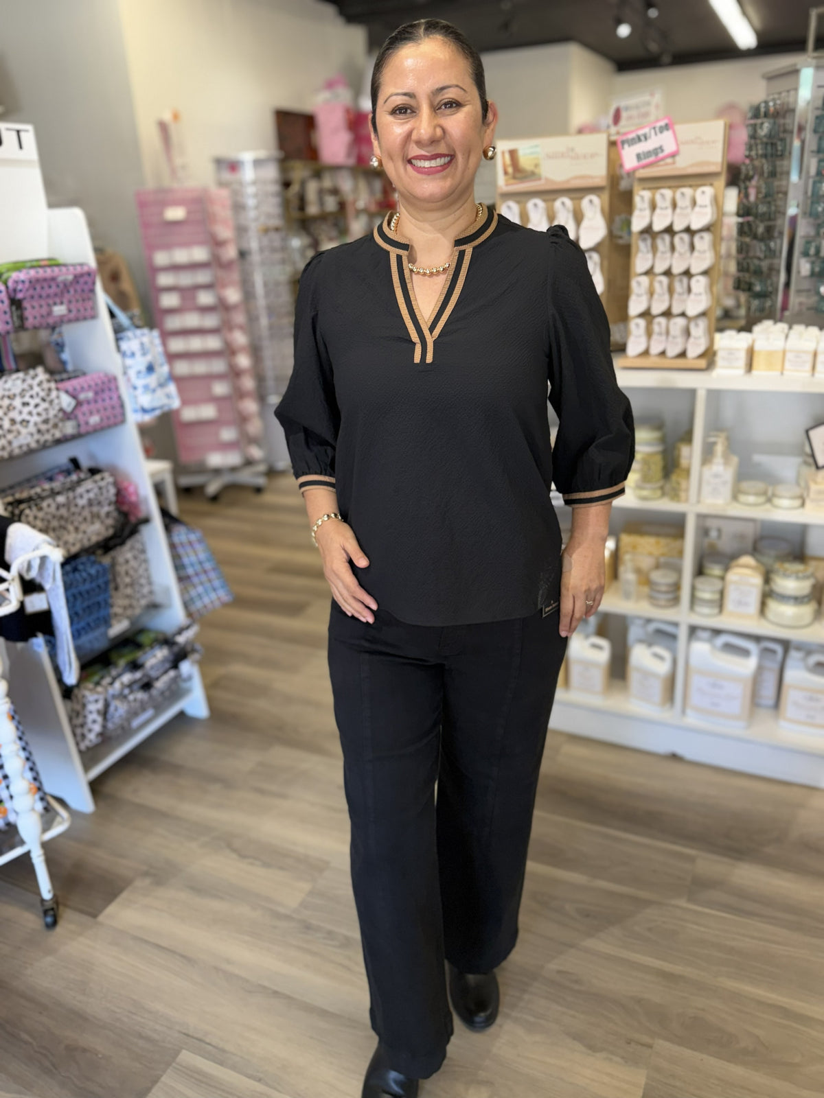 This is a classic black blouse with camel-tan contrast trim for a polished, sophisticated look. It features a split V-neckline with a short stand collar, accented by double tan piping that highlights the neckline. The three-quarter puff sleeves have gentle gathering at the shoulders and finish with coordinating striped cuffs. A relaxed, slightly flowy silhouette and subtle textured fabric make it an easy option for both work and dressier occasions.