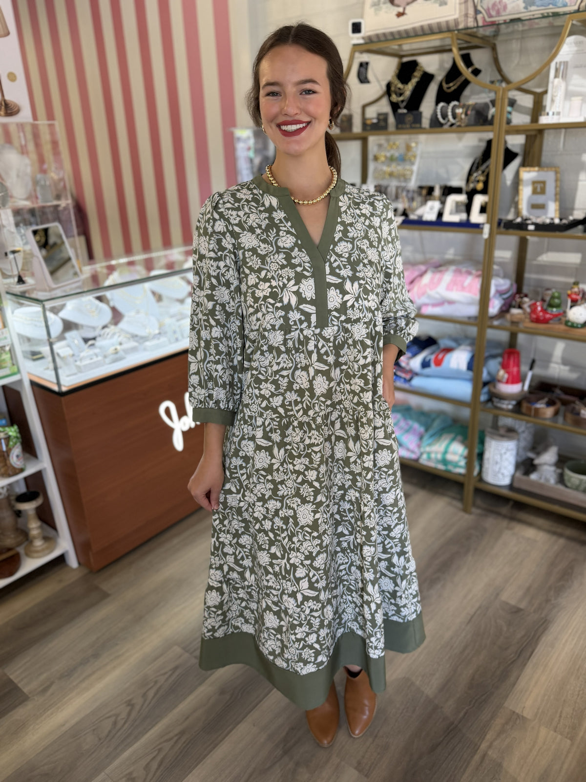 This is a sage green floral maxi dress featuring an allover white botanical print for a fresh, feminine look. It has a split V-neckline with solid sage trim, along with softly puffed three-quarter sleeves finished with coordinating cuffs. The relaxed bodice flows into a tiered, gathered skirt, creating an airy, flattering silhouette. A wide solid sage border at the hem adds a polished contrast and ties the design together. The flowy maxi length makes it a beautiful option for church, brunch, showers, family