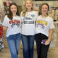 These are white collegiate graphic T-shirts featuring three North Carolina universities, each with a spirited, feminine design:

NC State Wolfpack Tee: Features “NC State University” above bold red WOLFPACK lettering with black accents.
UNC Tar Heels Tee: Features a vintage-inspired pennant graphic with TAR HEELS lettering, scattered stars, and the UNC logo in Carolina blue. Bold NORTH CAROLINA lettering runs underneath for a cla